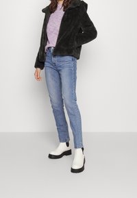 Chaqueta de pelo sintético negra con capucha, suéter de punto morado, jeans azul claro y botas de tobillo con tacón grueso blanco y acentos negros.