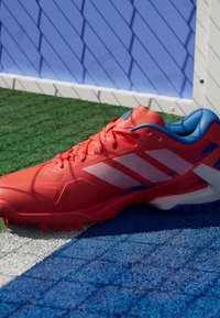 Red athletic shoe with white and blue accents, textured upper, and prominent laces. Features molded spikes for traction.
