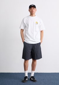 Hvit T-skjorte med korte ermer og gul grafikk, kombinert med mørke rutete shorts. Modellen har på seg hvite sokker og svarte joggesko.