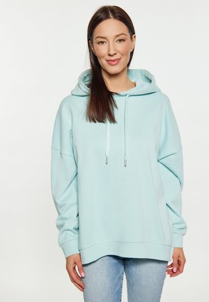 Junge Frau mit langen braunen Haaren lächelnd, trägt einen hellblauen Hoodie und hellblaue Jeans, steht vor einem weißen Hintergrund.