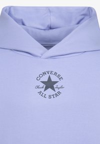 Világos lila kapucnis pulóver nagy szürke csillag logóval, amelyen a "CONVERSE" és az "ALL STAR" feliratok láthatók, sima szövet textúrával és húzózsinóros kapucnival.