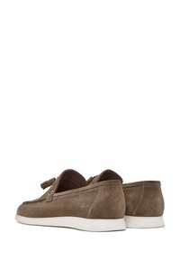 Mocasines de ante marrón con suela blanca plana, que presentan un acento de borlas y detalles de costura en la parte superior. Textura suave. Diseño casual.
