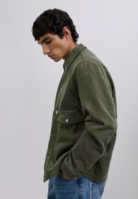 Joven en chaqueta de pana verde oliva y vaqueros azules, de pie de perfil con las manos en los bolsillos frente a un fondo sencillo.