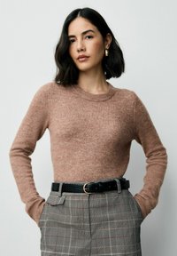 Sieviete ar īsiem melniem matiem, tērpta brūnā garām piedurknēm kreklā, raibās augstās jostas biksēs ar melnu jostu un zelta auskariem, stāv ar rokām kabatās.