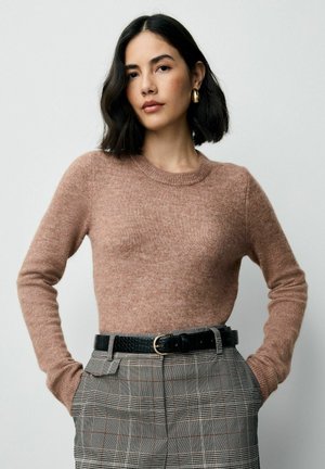 Frau mit kurzen schwarzen Haaren, die einen braunen Langarmpullover, karierte High-Waist-Hosen mit einem schwarzen Gürtel und goldenen Ohrringen trägt, und mit den Händen in den Taschen steht.