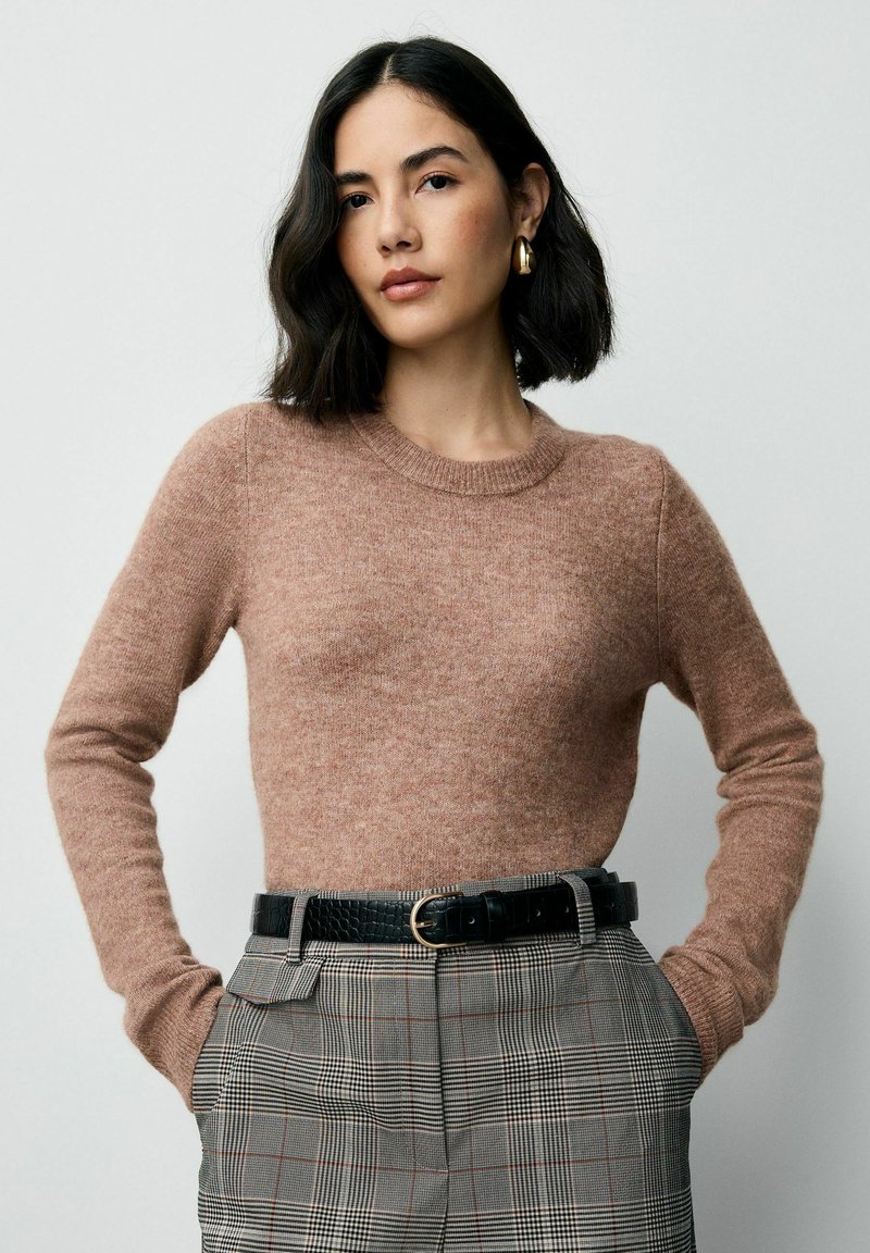 Sieviete ar īsiem melniem matiem, tērpta brūnā garām piedurknēm kreklā, raibās augstās jostas biksēs ar melnu jostu un zelta auskariem, stāv ar rokām kabatās.