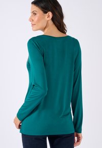 Langarmshirt in teal met een gladde textuur, ronde halslijn en subtiele versieringen. Het heeft een relaxed fit en reikt tot de heupen.