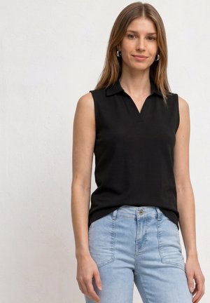 Frau mit langen hellbraunen Haaren, die ein ärmelloses schwarzes Oberteil und hellblaue High-Waist-Jeans trägt, steht an einer weißen Wand.