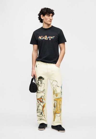 Μαύρο μπλουζάκι με πολύχρωμο γραφικό τύπωμα, κρεμ παντελόνι με καλλιτεχνικά σχέδια και μαύρα αθλητικά παπούτσια. Επιπλέον, μαύρη τσάντα στο χέρι.