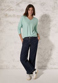 Maglione in tessuto turchese, scollatura a V, maniche tre quarti, abbinato a jeans a gamba larga blu scuro e sneakers bianche con dettagli neri.