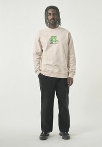 Beige sweatshirt with green graphic print and text, paired with black pants and black shoes. The fabric appears soft with a relaxed fit.