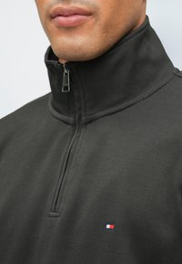 Felpa nera con zip e colletto alto, caratterizzata da un piccolo logo rosso, bianco e blu sul petto. Texture liscia e design minimalista.