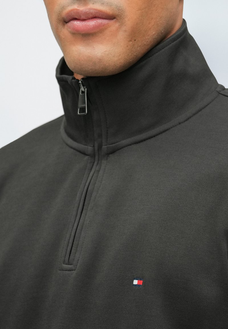 Felpa nera con zip e colletto alto, caratterizzata da un piccolo logo rosso, bianco e blu sul petto. Texture liscia e design minimalista.