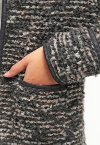 Texturerat svart och grått tyg med ett vågigt mönster; har en ficka med en hand placerad inuti; kontrasterande släta accenter vid kanterna.