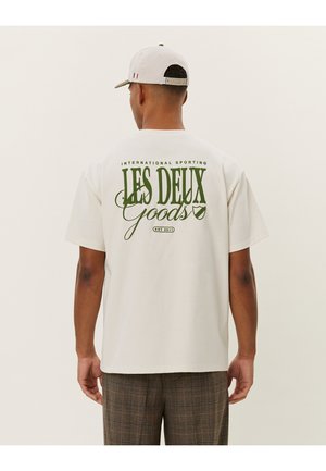 Camiseta blanca de manga corta con texto verde "LES DEUX Goods" y "INTERNATIONAL SPORTING" en la parte posterior; corte relajado y material de algodón.