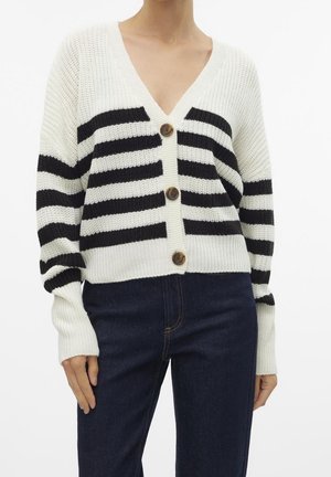 Femme portant un cardigan en maille blanc avec de larges rayures horizontales noires et de gros boutons marron, associé à un jean bleu foncé.