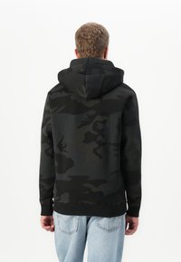 Hombre visto por detrás, vestido con una sudadera de capucha en camuflaje negro y gris oscuro, y jeans de color azul claro, contra un fondo blanco liso.