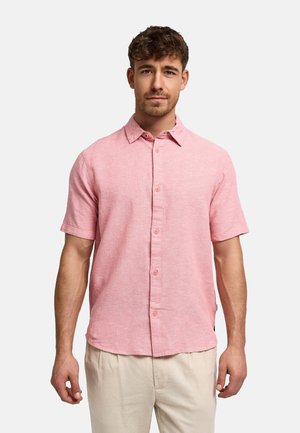 Nuori mies pukeutuneena vaaleanpunaiseen lyhythihaiseen napilliseen paitaan ja beigen värisiin housuihin, seisoo vaalean yksivärisen taustan edessä.