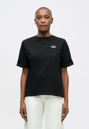 Person with shaved head wearing a black T-shirt with small white logo on chest and white pants, standing against plain white background.