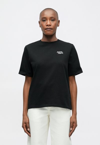 Άτομο με ξυρισμένο κεφάλι, φορώντας μαύρο T-shirt με μικρό λευκό λογότυπο στο στήθος και λευκά παντελόνια, σε στάση μπροστά από έναν απλό λευκό φόντο.