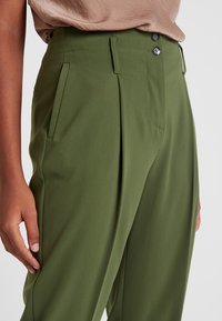 Pantalones color verde oliva con un corte entallado, pliegues y bolsillos frontales. El material parece suave con un ligero brillo. Cierre de dos botones.