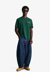 Green cotton t-shirt with a gold logo, paired with oversized blue denim jeans and brown work boots. Sunglasses worn on the face.