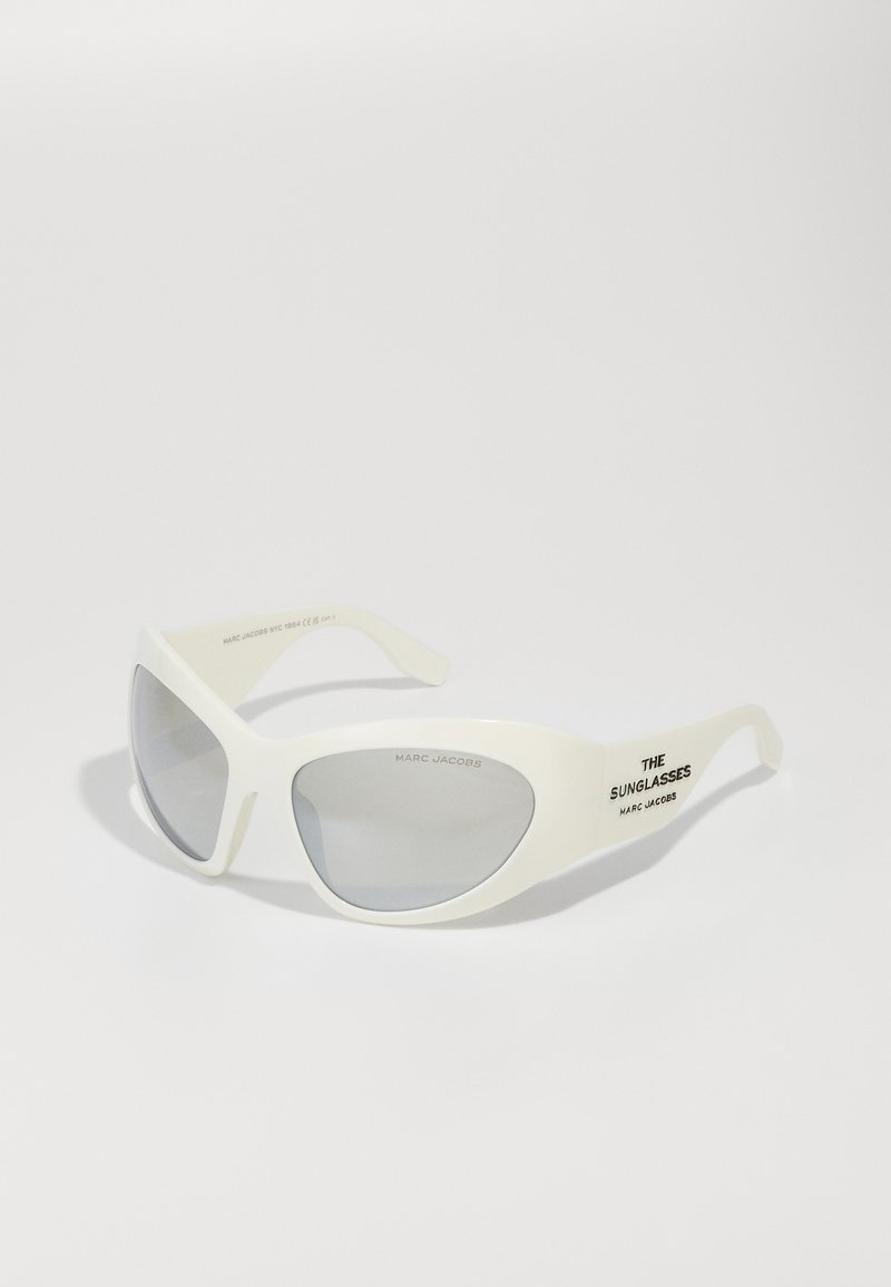 Valged plastikust päikeseprillid, millel on kaardus disain, hallid objektiivid ja must tekst "THE SUNGLASSES" külje peal.