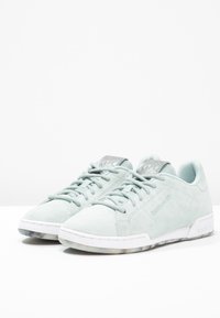 Ljusgröna mockasneakers med vita gummisulor, med en len textur, snörning och metalliska detaljer på tungans yta.