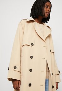 Béžový dvouřadý trench coat s velkými černými knoflíky, širokým límcem a ramenními epauletami; nošený přes pletený svetr.