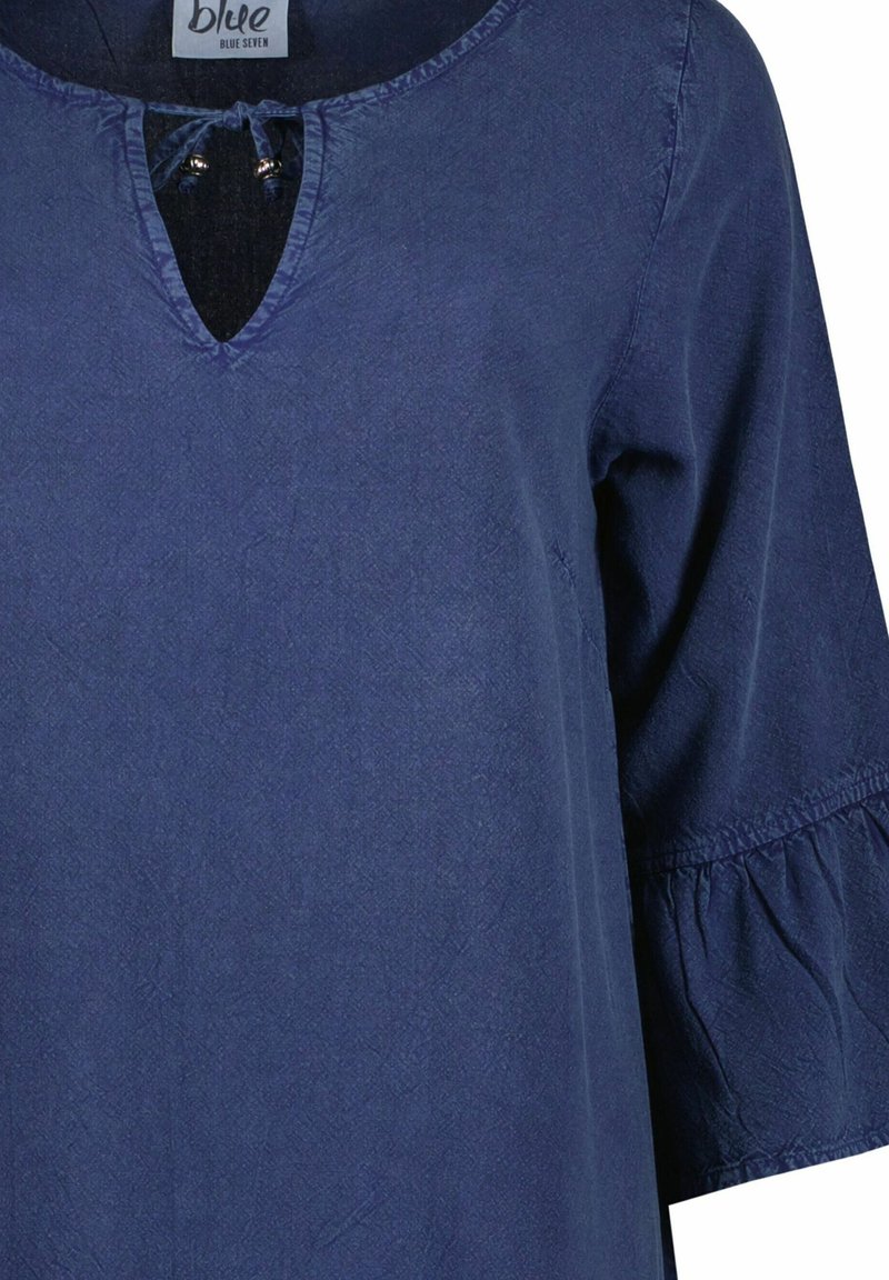 Marineblauwe tuniek met een v-hals en strikdetail, voorzien van driekwartmouwen met gerimpelde uiteinden, gemaakt van een gestructureerde stof.