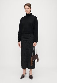 Melna adīta augstās apkakles džemperis, gara melna svārku ar smalku rakstu, brūna frizēta rokassoma un tumši brūni smaili apavi.