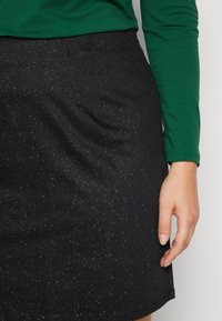 Jupe noire avec des paillettes scintillantes, faite d'un tissu doux. Coupe évasée avec un subtil éclat, associée à un long haut vert.