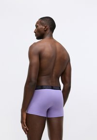 Lilla herre boxershorts med svart logobånd, glatt tekstur og tettsittende design, vist fra baksiden mot en ensfarget bakgrunn.