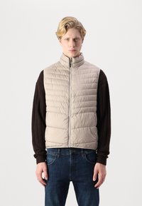 Beige quilted vest with a high collar, zippered front, and dark brown long sleeves. Worn over blue denim jeans.