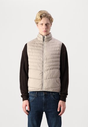 Beige quilted vest with a high collar, zippered front, and dark brown long sleeves. Worn over blue denim jeans.