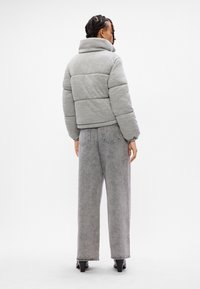 Kvinde står med ryggen til, iført en grå polstret jakke, grå brede bukser og sorte hælede sko mod en hvid baggrund.