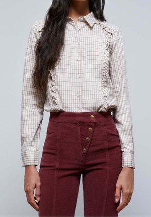 Vrouw draagt een wit geruit overhemd met knopen en ruches, en een hooggetailleerde bordeauxrode broek met gouden knopen, staand tegen een effen achtergrond.