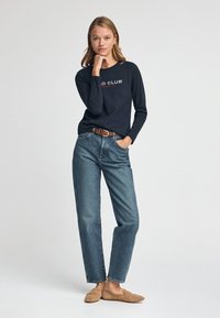 Camicia a maniche lunghe blu navy con logo bianco e rosso, abbinata a jeans azzurri a vita alta e scarpe beige slip-on. Outfit semplice e casual.