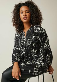 Blusa nera con motivi astratti bianchi, fronte plissettato e maniche lunghe a palloncino. Tessuto morbido con una vestibilità comoda e un leggero lucido.