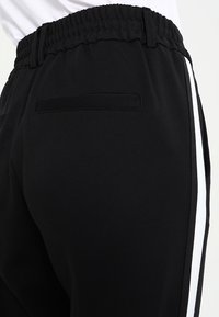 Svarta idrottsbyxor i mjukt material, med en vit sidremsa och resårmidja. Inkluderar en liten bakficka.