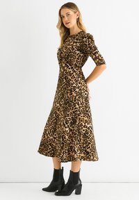 Leopardenmusterkleid in Tan und Schwarz, mit einem taillierten Oberteil, kurzen Ärmeln und einem ausgestellten Midirock. Kombiniert mit schwarzen Stiefeletten.