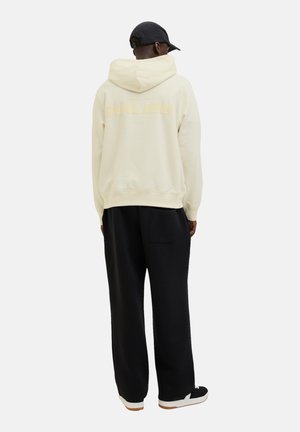 Lys beige hættetrøje med "MALIBU" trykt på ryggen, parret med løstsiddende sorte bukser og sorte sneakers.