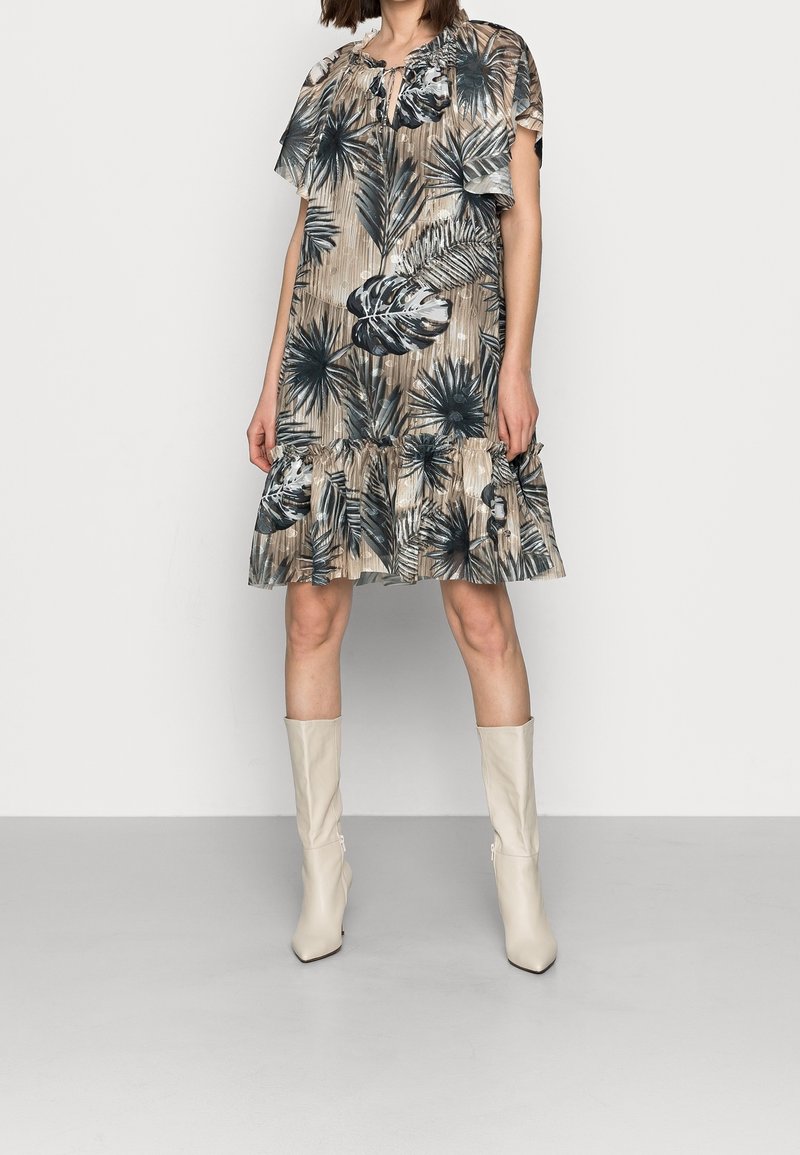 Femme portant une robe beige mi-longue avec des imprimés de feuilles tropicales noir et blanc et des bottines beige mi-mollet, debout devant un fond uni.