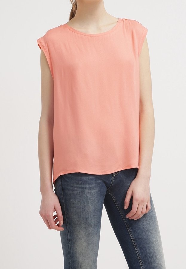 Femme portant un chemisier corail rose sans manches et un jean bleu délavé, debout devant un fond clair uni.
