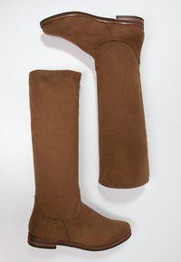 Bottes hauteur genou en daim marron avec une texture lisse, un embout arrondi et un talon plat. Caractéristiques : coutures surpiquées et tige ajustée.