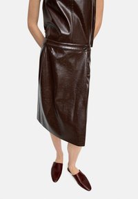 Brąsowa skórzana sukienka o strukturalnym kroju, z detalem w postaci guzika z boku i miękką teksturą. Połączona z ciemnobrązowymi butami wsuwanymi.
