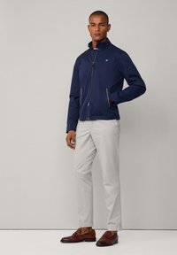 Chaqueta de zip azul marino con cuello alto, dos bolsillos delanteros con ribete marrón, combinada con pantalones gris claro y mocasines marrones.