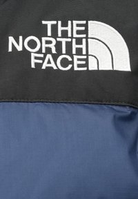 Crna i mornarsko plava jakna s logotipom "THE NORTH FACE" u bijeloj vezenoj izvedbi na gornjem dijelu, s glatkom teksturom tkanine otpornom na vodu.