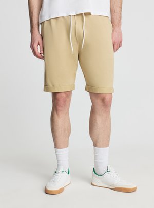 Mężczyzna ubrany w beżowe krótkie spodenki z podwiniętymi brzegami, białe skarpetki za kostkę, białe tenisówki z zielonymi akcentami oraz białą koszulę, stojący na jednolitym tle.