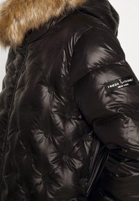 Chaqueta acolchada negra con capucha de piel sintética y estampado del símbolo de la paz. Cuenta con un cierre de cremallera y una etiqueta negra en la manga.